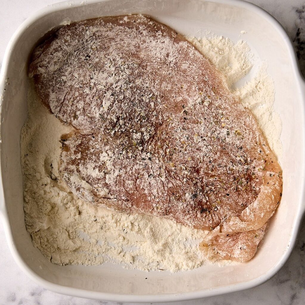 Thin chicken cutlets dusted with flour and Italian seasoning in a white dish, loose flour scattered around them.