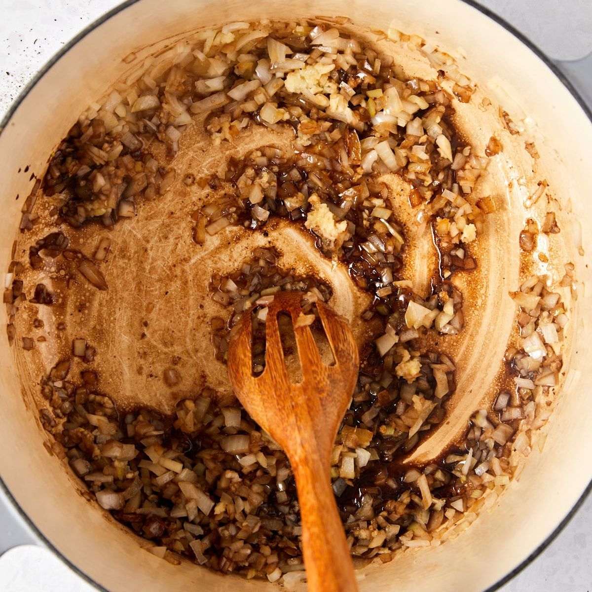 A wooden spoon stirs sautéed onions and garlic browning in a light-colored Dutch oven.