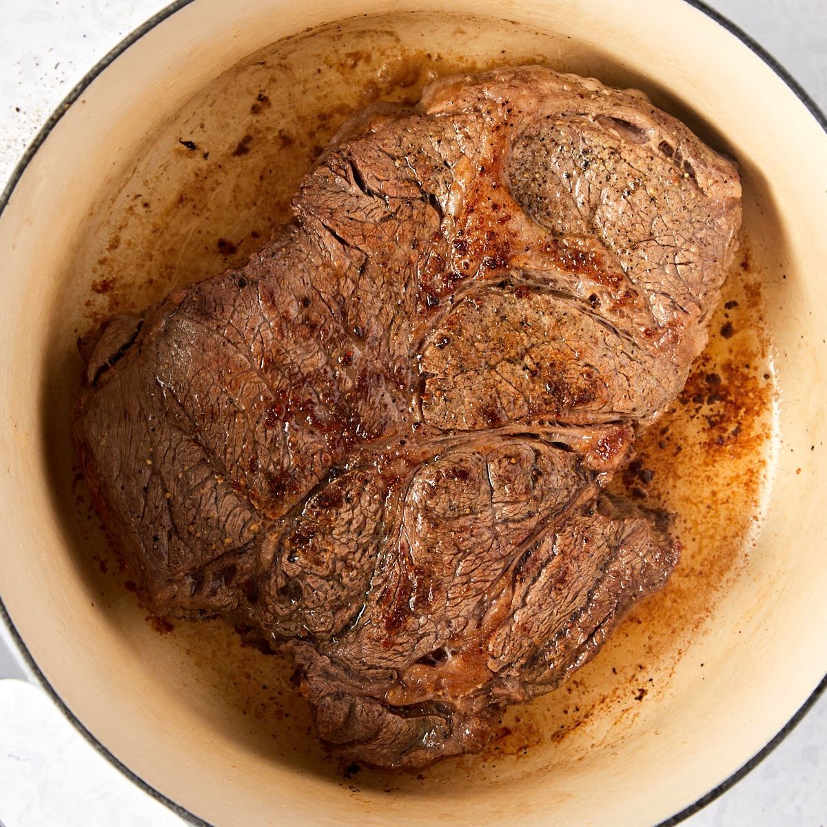 A large, browned beef roast searing in a light-colored enameled cast iron pot.