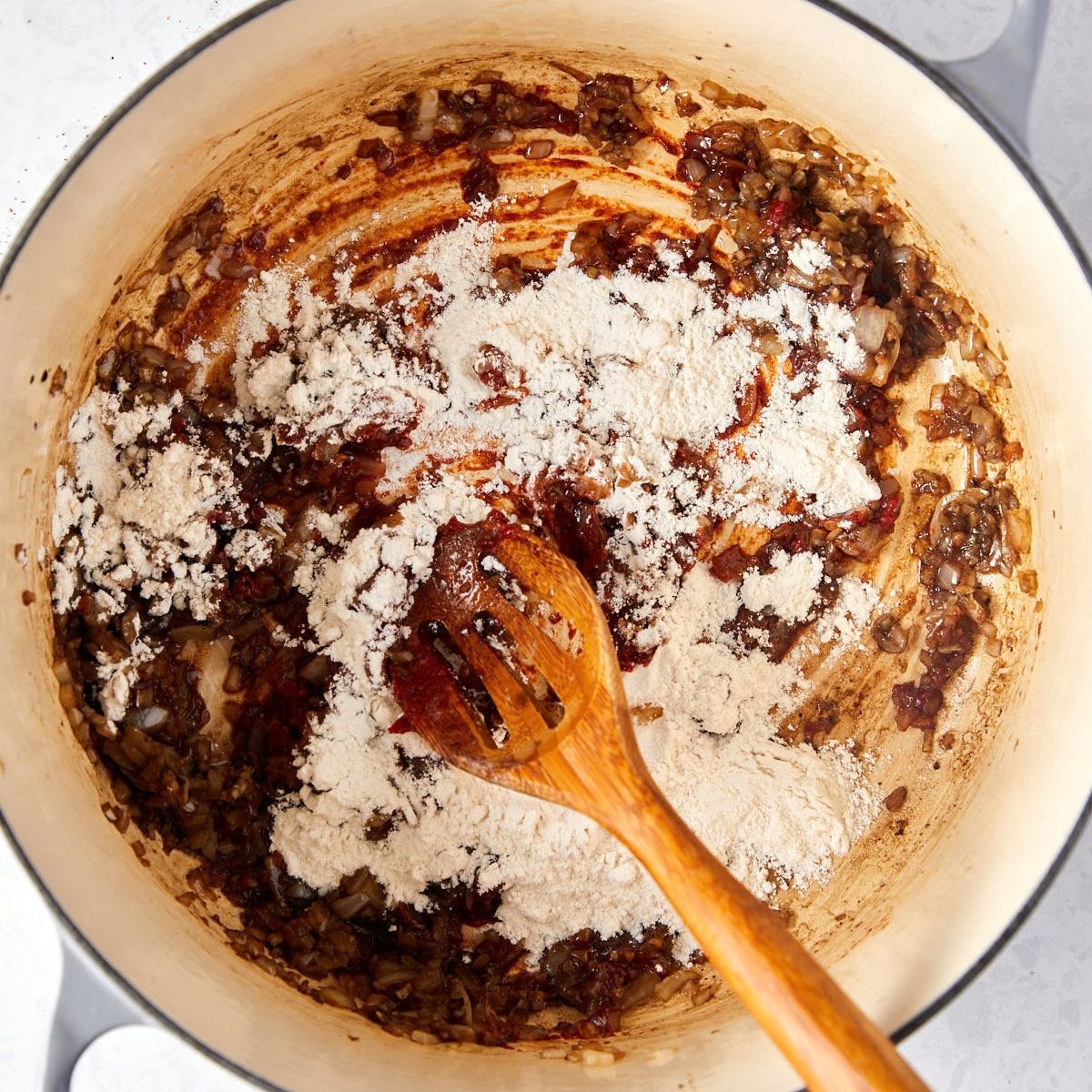 A wooden spoon stirring a mound of white flour into sautéed onions and tomato paste in a large enameled pot.