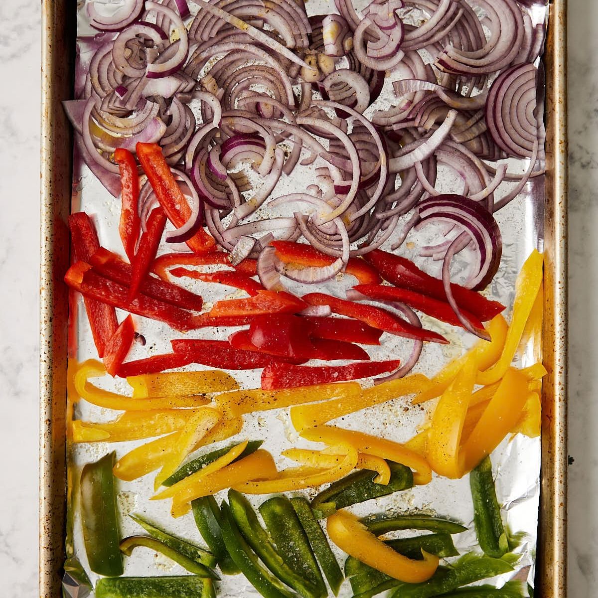 Sliced red onion and red, yellow, and green bell peppers on a foil-lined sheet pan, ready to roast.