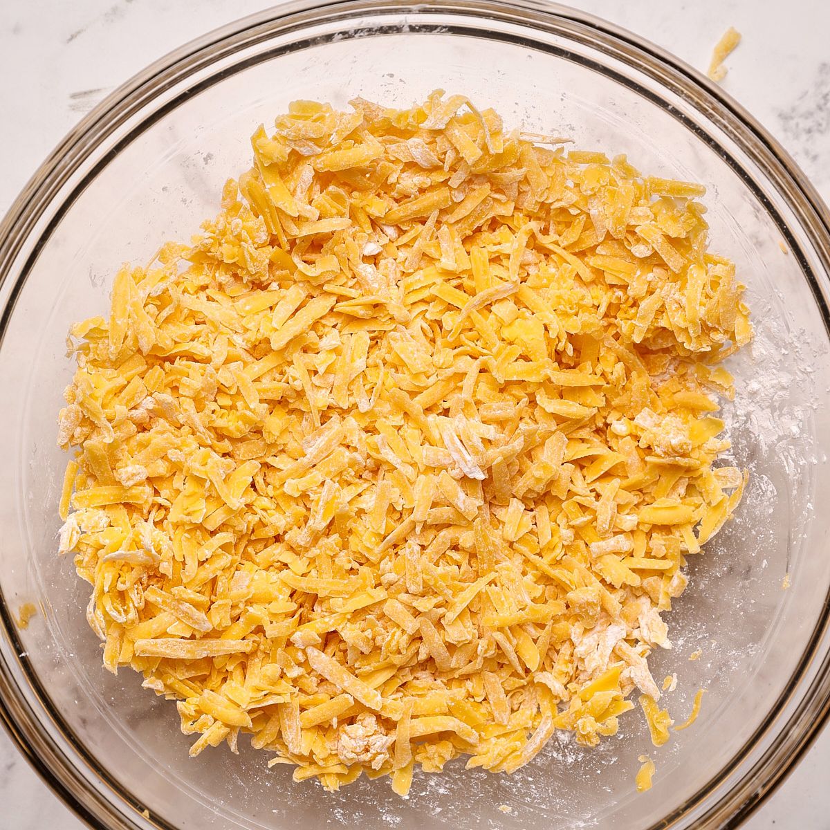 Shredded cheddar cheese tossed with cornstarch in a glass bowl.