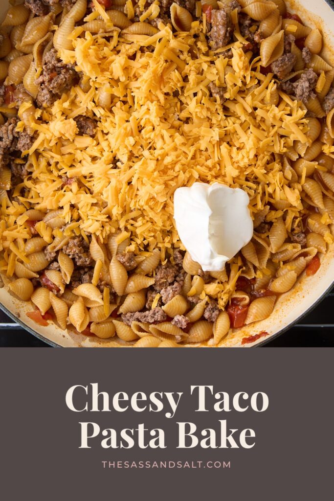 A skillet of taco pasta topped with shredded cheddar cheese and a dollop of sour cream, with text below reading “Cheesy Taco Pasta Bake” and the website URL.