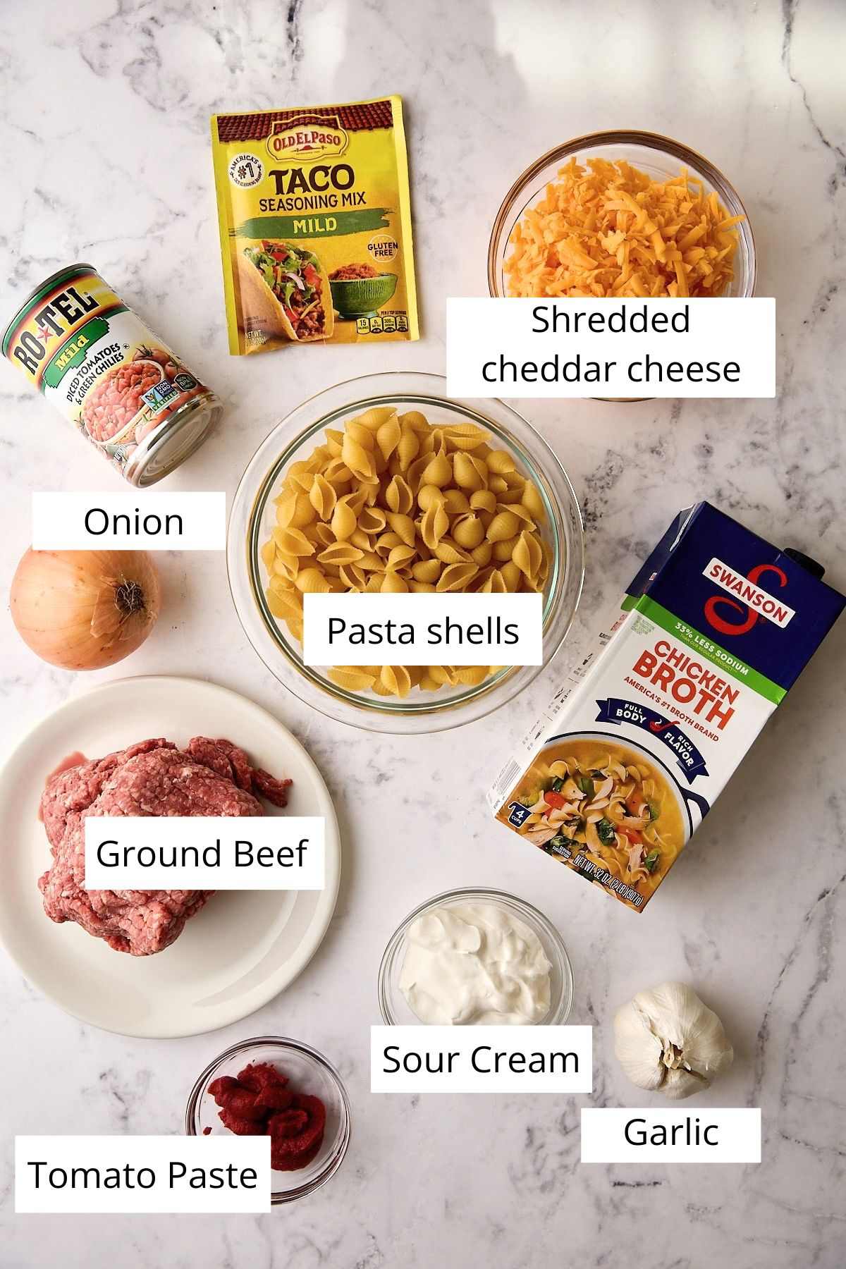 Ingredients for one-pot taco pasta: ground beef, medium pasta shells, block cheddar, taco seasoning, Rotel, chicken broth, onion, garlic, tomato paste, and sour cream.