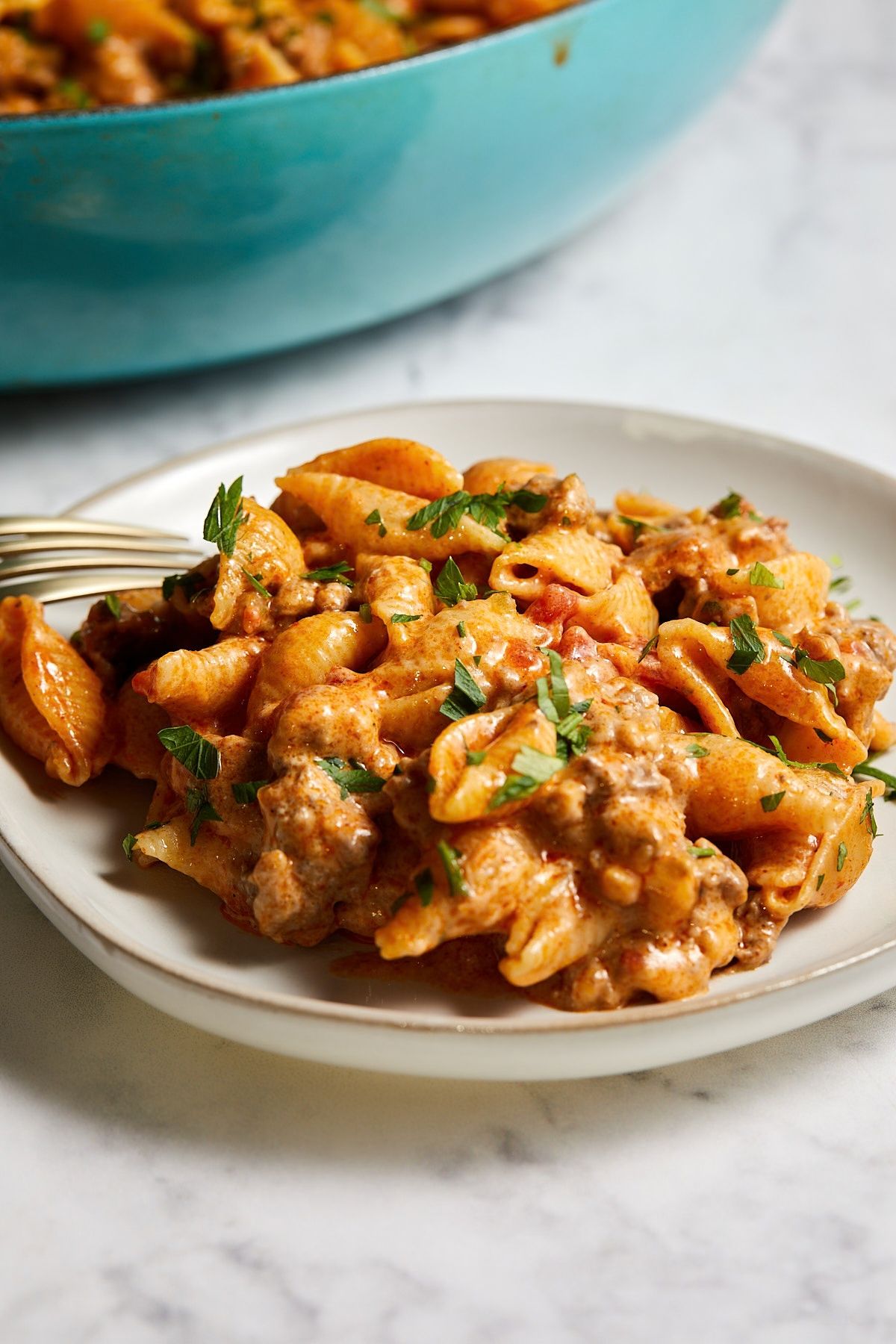 Taco pasta shells with ground beef in a creamy cheddar sauce on a white plate, with the blue braiser in the background.